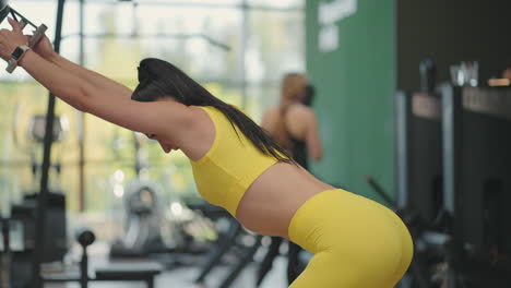 A-young-Hispanic-woman-in-a-yellow-tracksuit-performs-an-exercise-in-a-crossover-pulls-a-steel-rope-from-above-to-train-her-back-and-shoulders.-A-woman-trains-her-back-and-shoulders-in-a-gymnasium